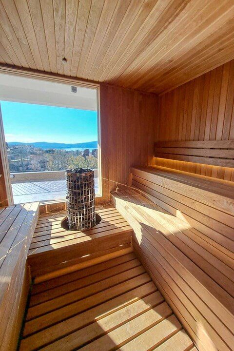 Modern Finnish sauna interior with light-colored wood benches and a panoramic view of the Helsinki seaside through a large glass window.