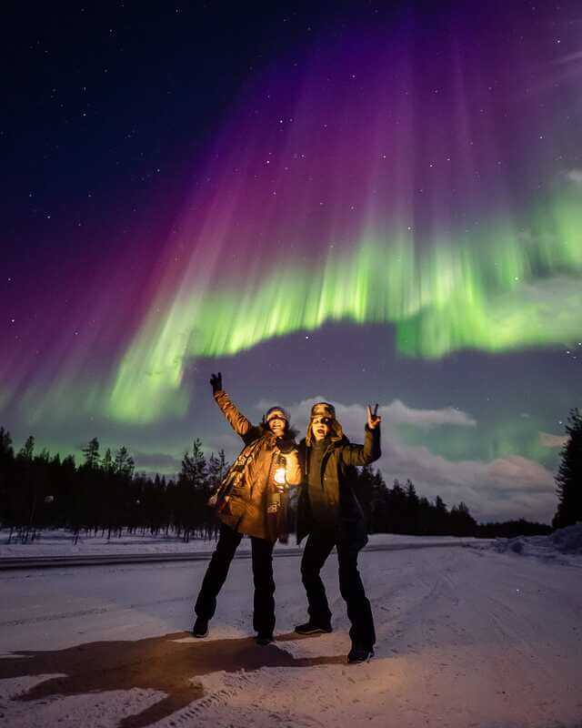 2 people with Stunning Northern Lights (Aurora Borealis) illuminating the night sky over a snow-covered forest and cabins in Finland.