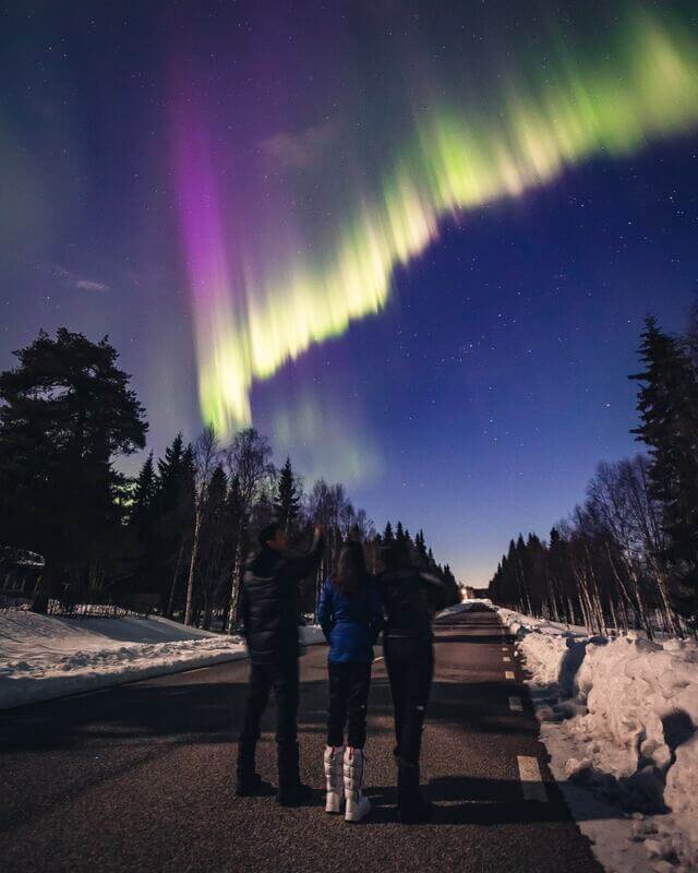 3 people with Stunning Northern Lights (Aurora Borealis) illuminating the night sky over a snow-covered forest and cabins in Finland.