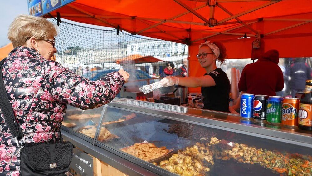 A friendly vendor serving fresh street food at a vibrant orange stall in Kauppatori Market, Helsinki.