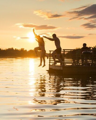 Silhouettes of people jumping off a wooden pier into a lake during a vibrant sunset, capturing the spirit of summer fun and the authentic Finnish "mökki" (cottage) lifestyle.