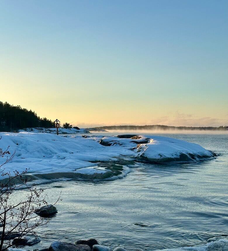 Ethereal mist rising from the calm Baltic Sea along the snow-covered rocky coastline of the Finnish archipelago during a serene winter sunrise.