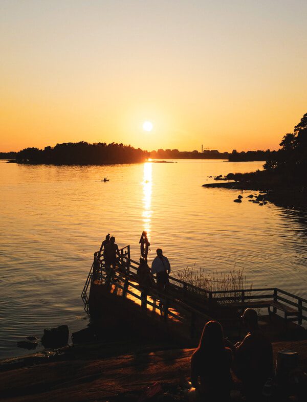 Silhouettes of people on a wooden pier overlooking the calm Baltic Sea at sunset, capturing the quintessential "Finnish joy" (ilo) of a summer evening.