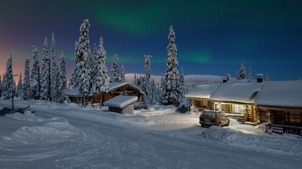 Stunning green Northern Lights (Aurora Borealis) illuminating the night sky over snow-covered log cabins in a Finnish winter landscape.